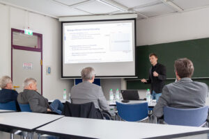 Mann stehend beim Vortrag vor 4 sitzenden Männern am Tisch