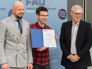 Drei Personen, wovon der in der Mitte eine Auszeichung in der Hand hält