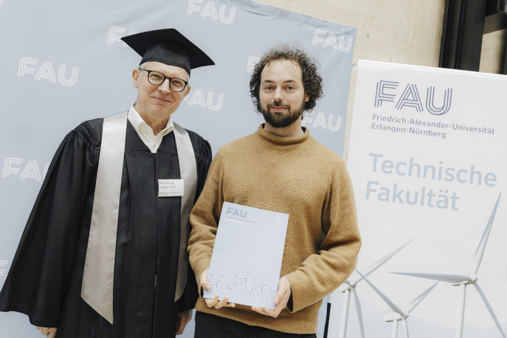 Zwei Personen vor einen Poster, eine Person hält ein Zeugnis in der Hand